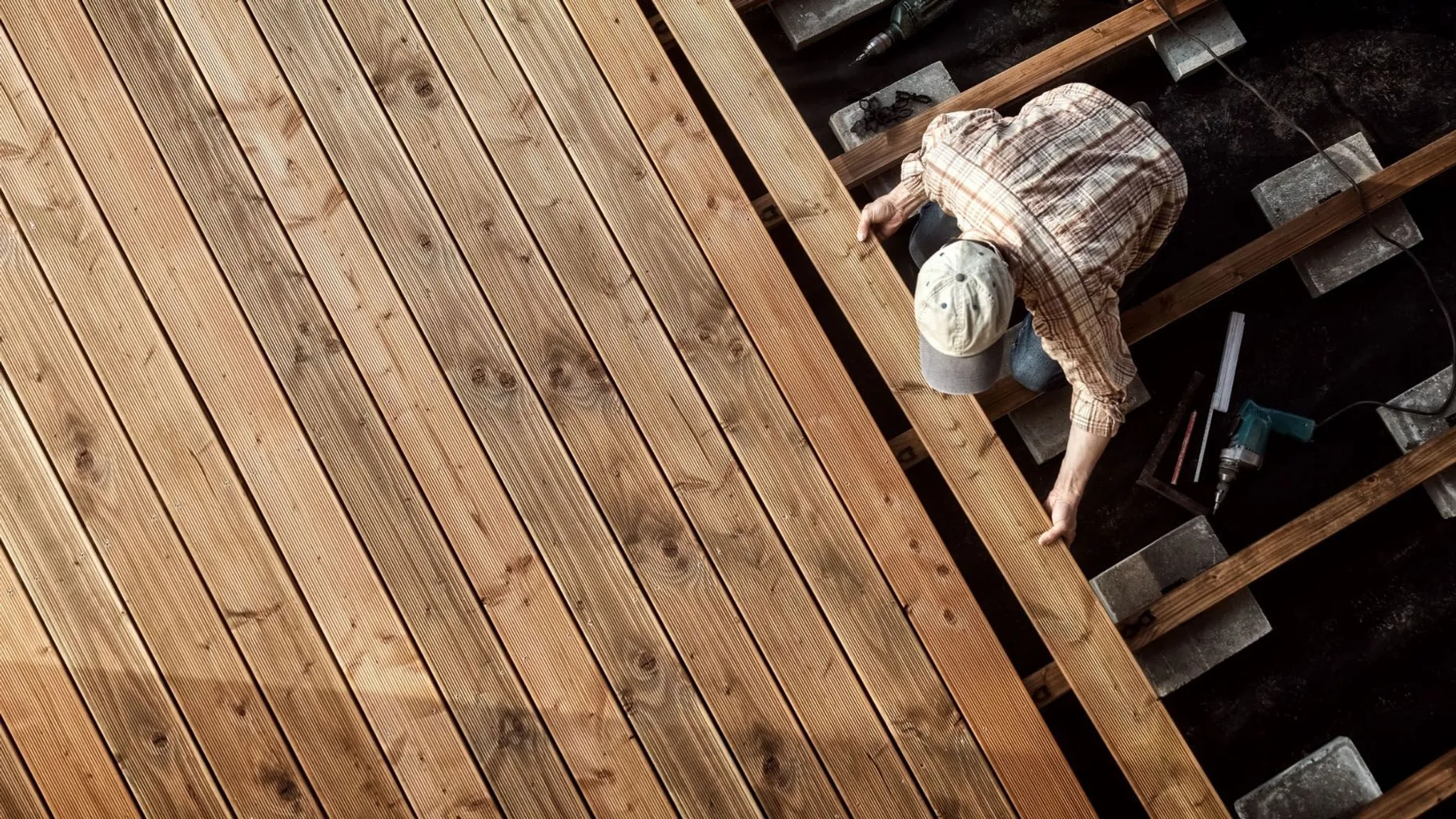 Eine Person verlegt braune Terrassendielen auf einer Unterkonstruktion mit Betonauflagen und Werkzeug daneben